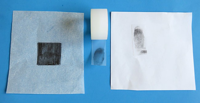 A paper square with a black square at its center next to a roll of tape and another paper square with a black fingerprint